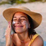 Woman applying sunscreen to her face while wearing a beach hat.