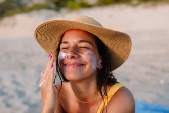 Woman applying sunscreen to her face while wearing a beach hat.